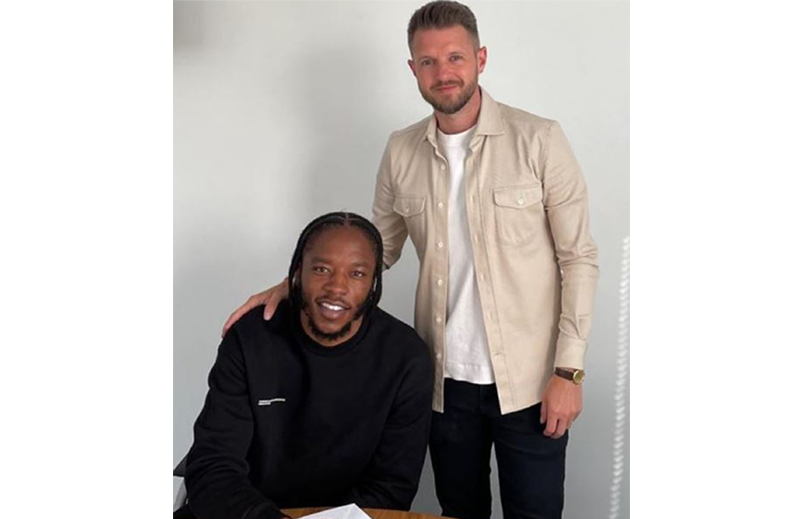 Romaine is sat at a table signing a contract, Scott is stood next to him with his hand on Romaine's shoulder. They are both smiling, looking happy