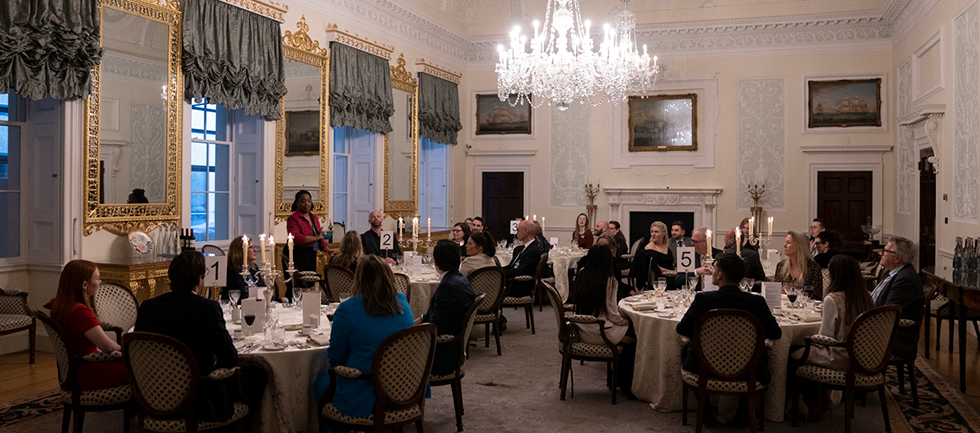 The group is sat and round dinner tables in the Adam room watching someone stood up and speaking to the room