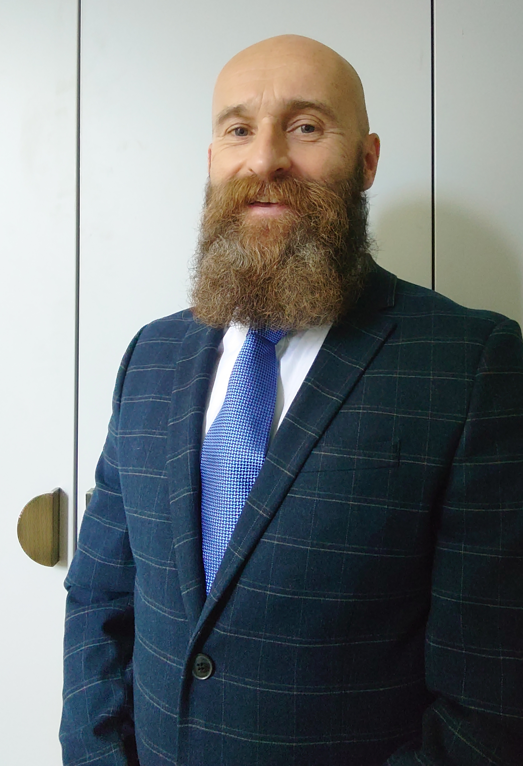 Mark has a beard, is smiling and is wearing a navy checked shirt jacket, a white shirt and a mid-blue tie