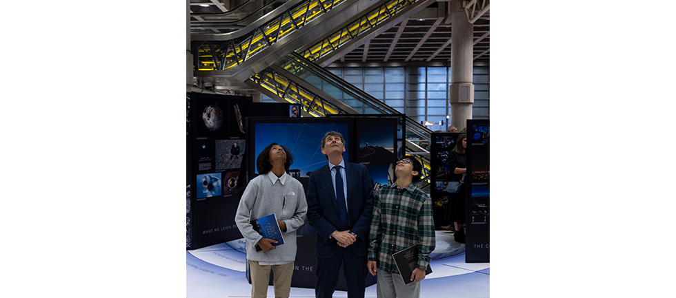 competition winners and max alexander standing next to each other look up to sky, they are standing in front of the exhibition with a background of the escalators in the Lloyd's underwriting room