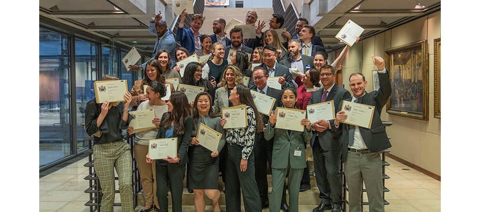 group standing on stairs, celebrating with their certificates. Each person is standing in a different pose, some people are cheering with their hands in the air, some are waving their certificates in the air, others are looking at each other and laughing. 